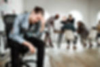 Young man in the foreground sitting on an office chair clutching his head in pain, with a boardroom or working employees in the background