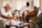 Senior living facility common room with various residents seated. A male is playing chss with an unseen man, a woman is knitting on the couch while a Doctor takes the blood pressure of a female in a rocking chair